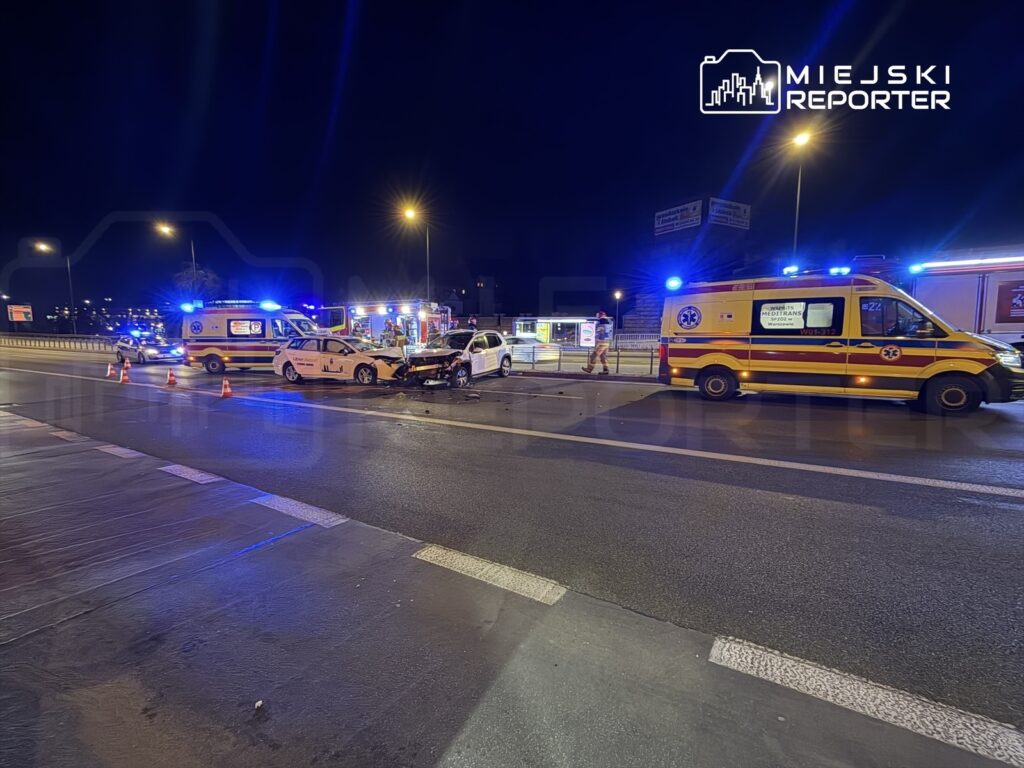 Na drodze nocą doszło do wypadku, w którym uczestniczyły dwa samochody. Obok stoją karetki pogotowia oraz radiowóz, a teren otoczony jest pachołkami.
