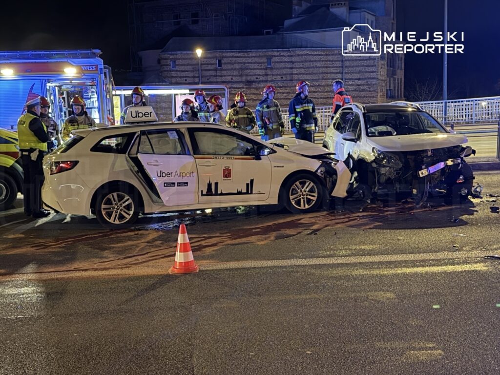 W nocy na ulicy doszło do zderzenia dwóch samochodów, w tym pojazdu Uber, z widocznymi uszkodzeniami. Obok pracują strażacy i policjanci.