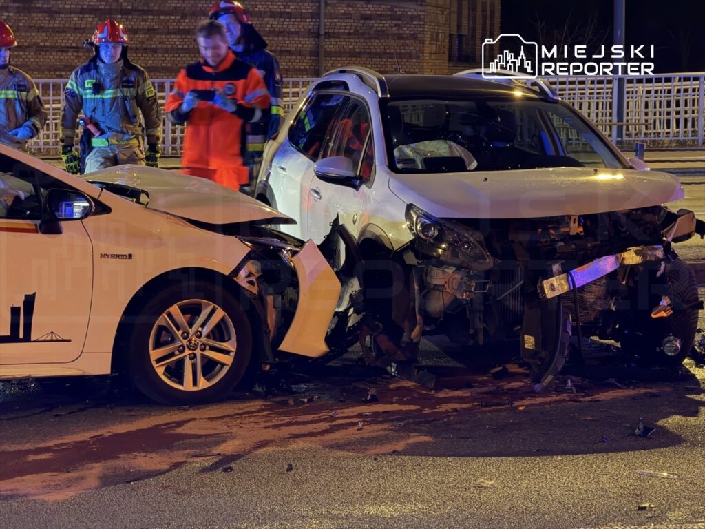Dwa uszkodzone samochody po zderzeniu, w tle strażacy i ratownicy medyczni oceniają sytuację na nocnej ulicy.