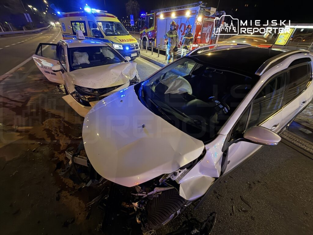 Dwa uszkodzone samochody osobowe stoją na drodze, obok nich pojazdy ratunkowe z włączonymi sygnałami świetlnymi.
