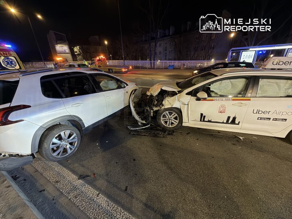 Dwa uszkodzone pojazdy po zderzeniu na ulicy, w tle widać pojazdy służb ratunkowych i oświetlenie nocne.