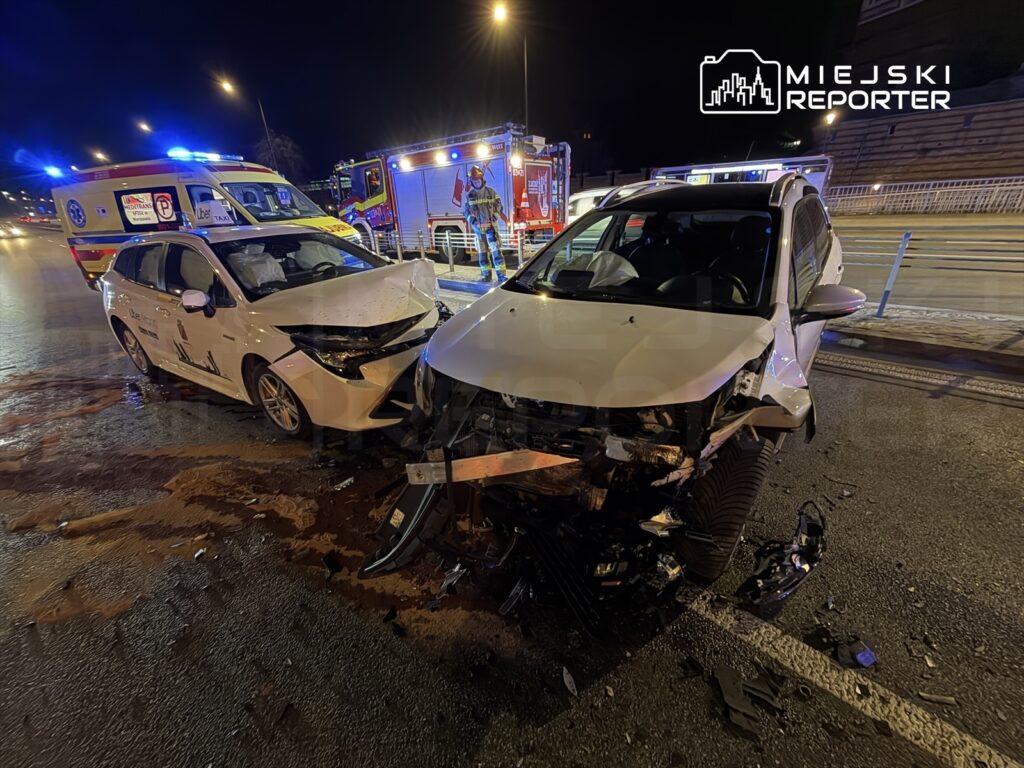 Dwa uszkodzone samochody osobowe po kolizji na drodze, w tle widać karetki pogotowia i służby ratunkowe.