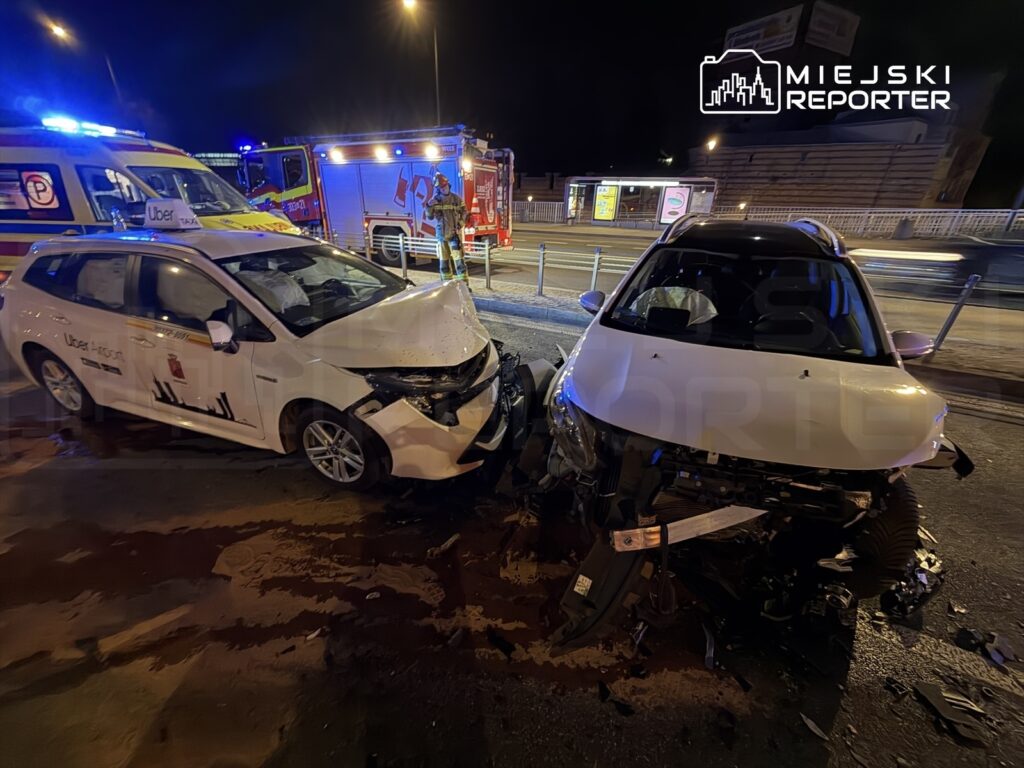 Dwa uszkodzone samochody osobowe po wypadku drogowym na miejskiej ulicy, w tle widać pojazdy służb ratunkowych.