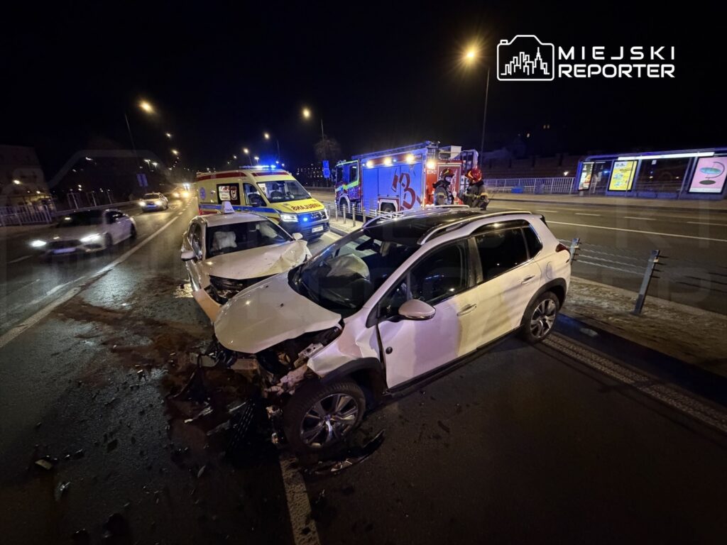 Dwa rozbite samochody na jezdni, obok stoją karetka i wóz strażacki, w tle nocne oświetlenie ulicy.