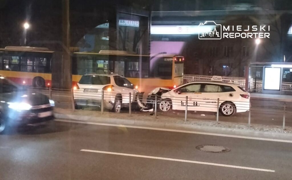 Dwa uszkodzone samochody stoją obok przystanku tramwajowego, w tle widoczny tramwaj i nocne oświetlenie ulicy.