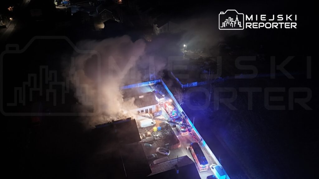 Pożar w budynku, który otoczony jest gęstym dymem, z widocznymi wozami strażackimi i ratownikami w akcji.