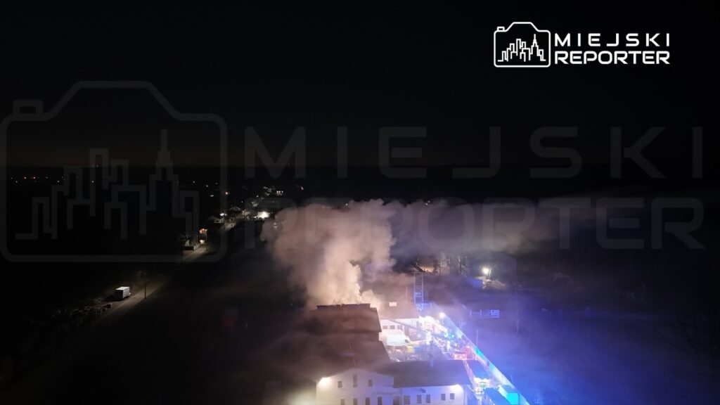 W nocy dym unosi się nad budynkami, a w tle widoczne są oświetlone ulice i pojazdy.