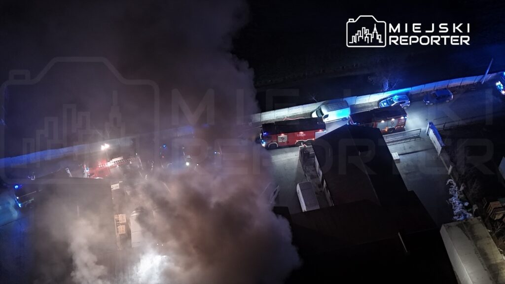 Strażacy w akcji gaszenia pożaru, otoczeni gęstym dymem, przy budynku z widocznymi pojazdami ratunkowymi.
