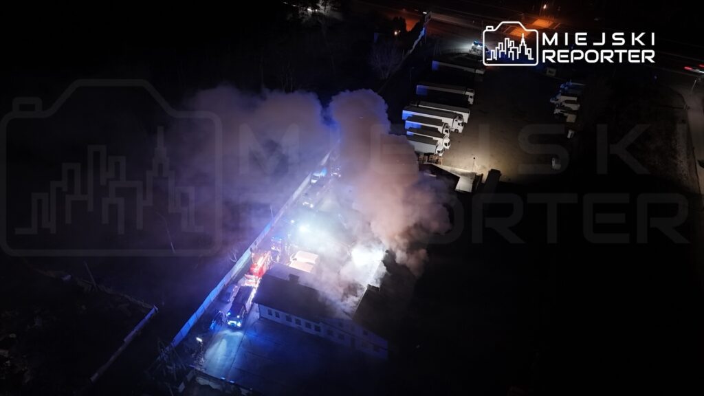 Gęsty dym unoszący się nad budynkiem, obok którego stoją pojazdy straży pożarnej, w nocy.
