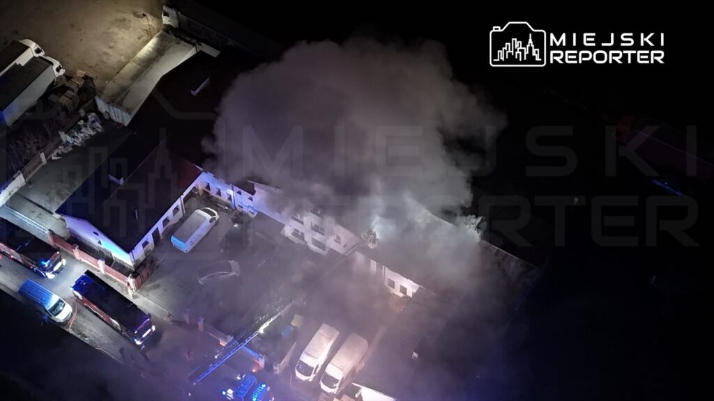 Gęsty dym wydobywający się z budynku, w otoczeniu pojazdów straży pożarnej oraz oświetlenia akcji ratunkowej.