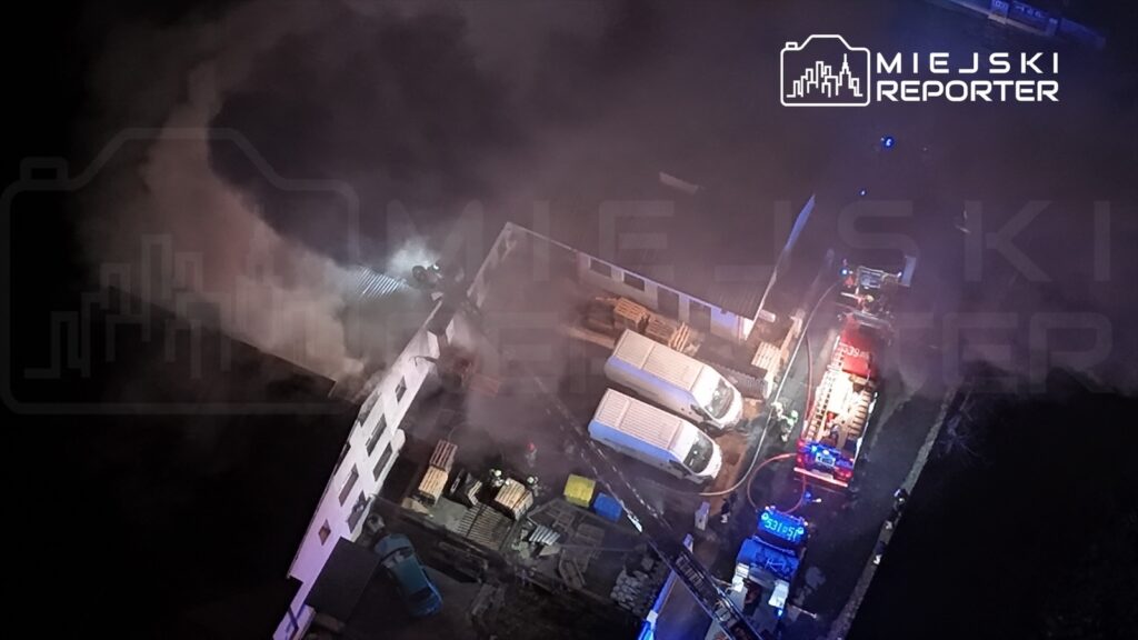 Dym wydobywający się z dachu budynku, w pobliżu stojące samochody strażackie oraz ratownicy na miejscu akcji.