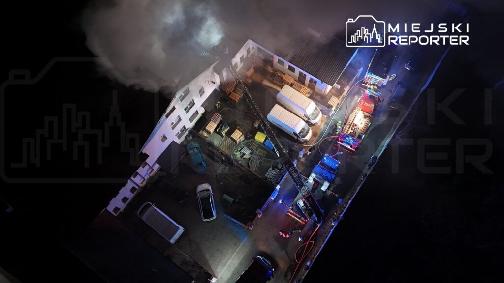 Strażacy gaszą pożar w budynku przemysłowym, otoczeni przez pojazdy ratunkowe i unoszący się dym.