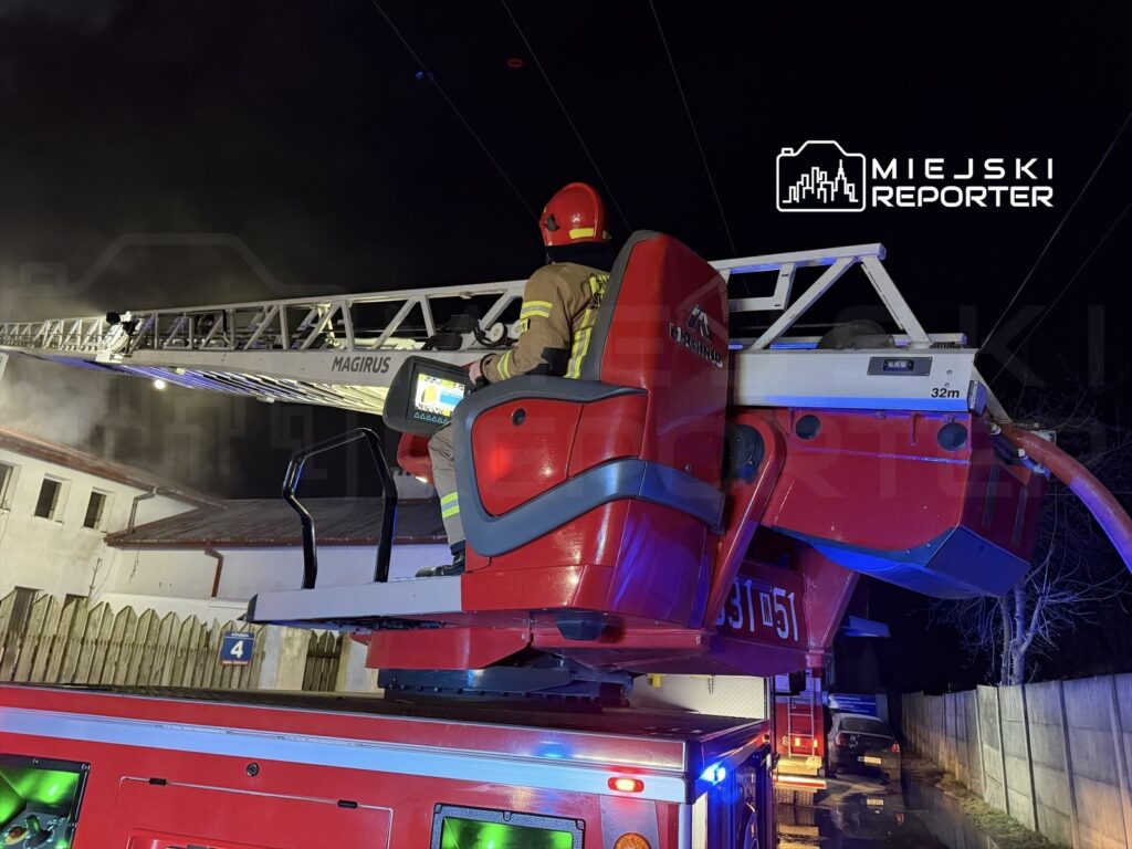 Strażak w hełmie siedzi na podnośniku strażackim, podnosząc drabinę w kierunku dymu nad budynkiem nocą.
