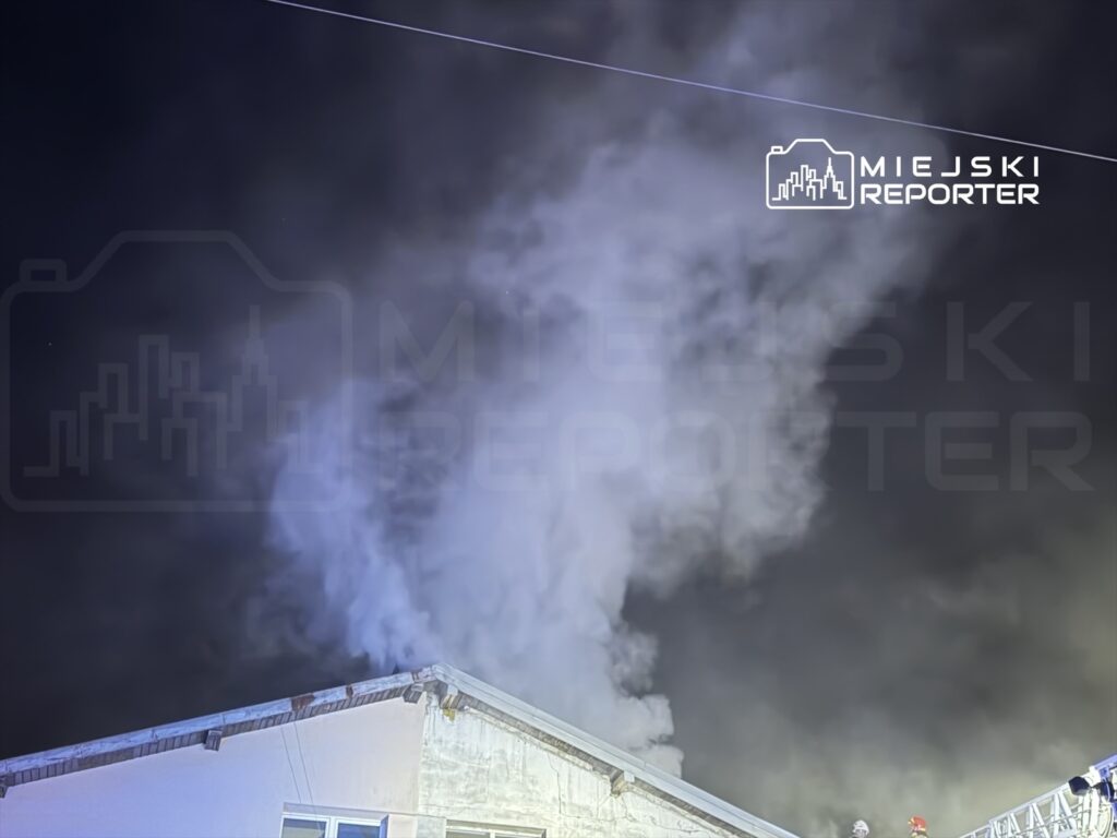 Gęsty dym wydobywający się z dachu budynku w nocy, z widocznymi strażakami w hełmach na szczycie.