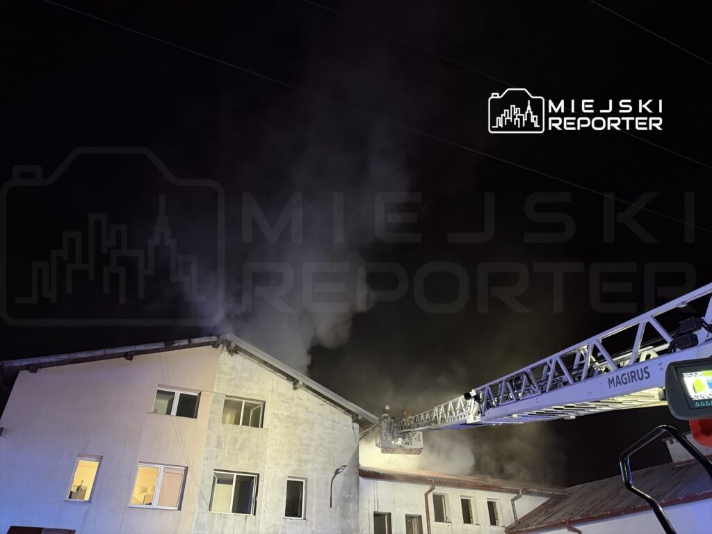 Strażacy na długiej drabinie gaszą pożar dachu budynku w nocy, z którego wydobywa się dym.