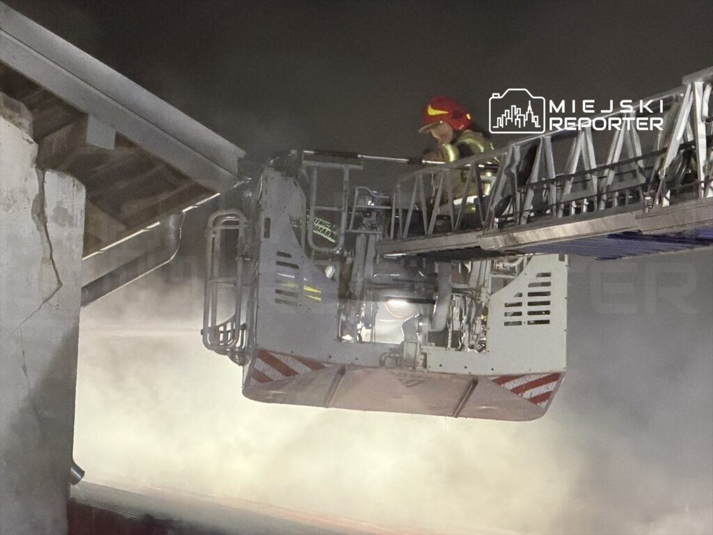 Strażak w kasku i kamizelce ochronnej obsługuje podnośnik strażacki w otoczeniu dymu.