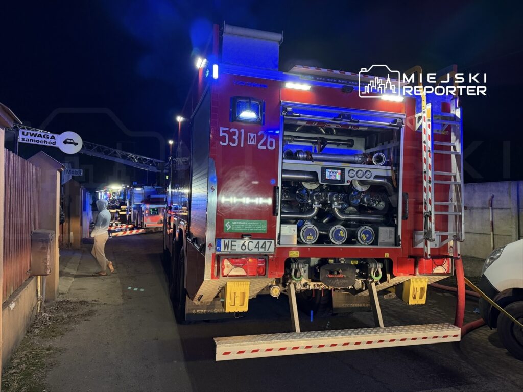 Straż pożarna z włączonymi sygnałami świetlnymi stoi na wąskiej ulicy, a obok niej widoczna osoba w jasnym ubraniu.