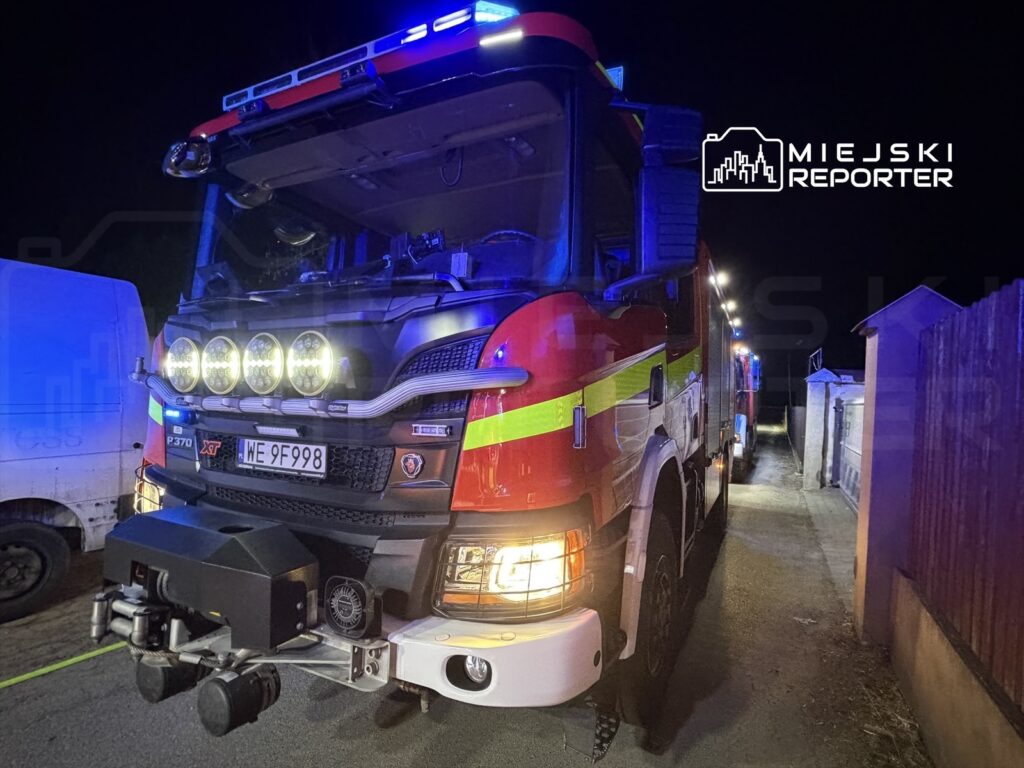 Straż pożarna z niebieskimi sygnałami świetlnymi stoi na wąskiej ulicy nocą, obok białego furgonu.