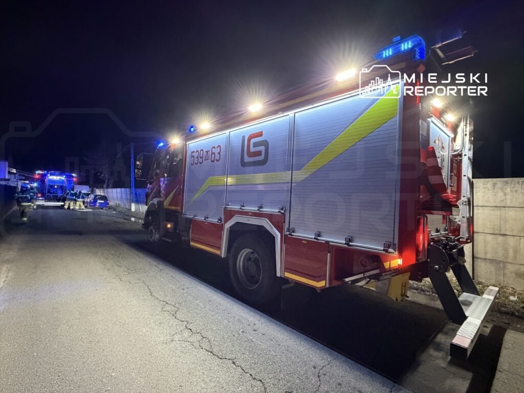 W nocy na ulicy zaparkowany wóz strażacki z włączonymi sygnałami świetlnymi, obok zespół strażaków interweniuje.