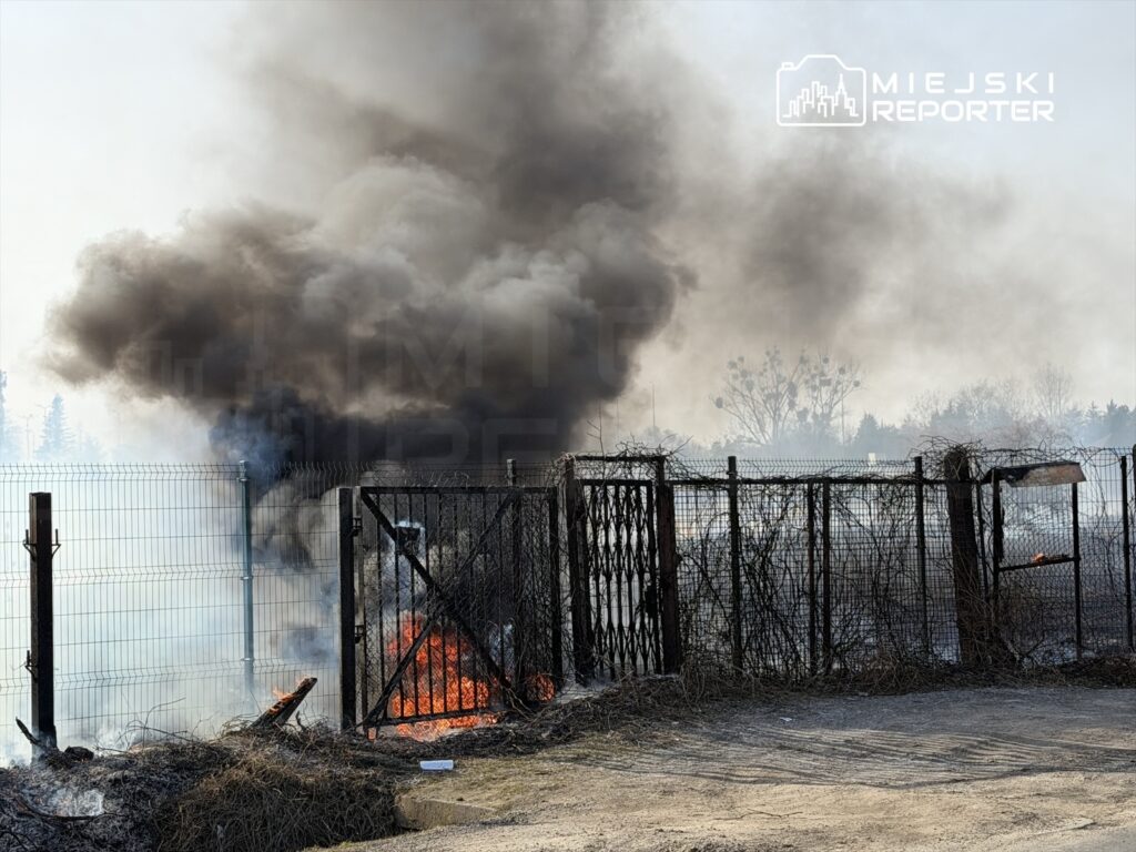 Wielka chmura czarnego dymu unosząca się nad płonącym ogniskiem w zaroślach przy ogrodzeniu.