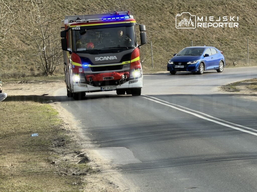 Wóz strażacki z włączonymi sygnałami świetlnymi skręca w prawo na skrzyżowaniu, obok siedzi mężczyzna z telefonem.