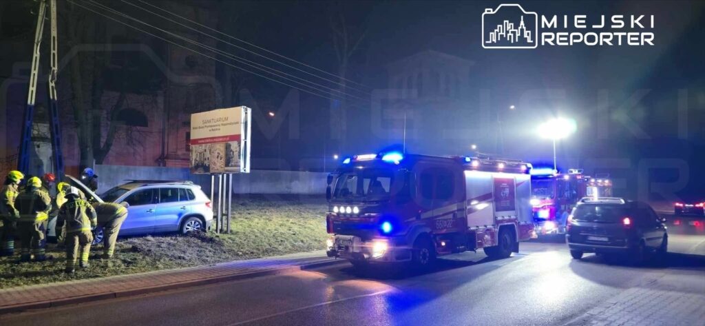 Grupa strażaków pracuje nad usunięciem uszkodzonego samochodu zaparkowanego na poboczu drogi w nocy.