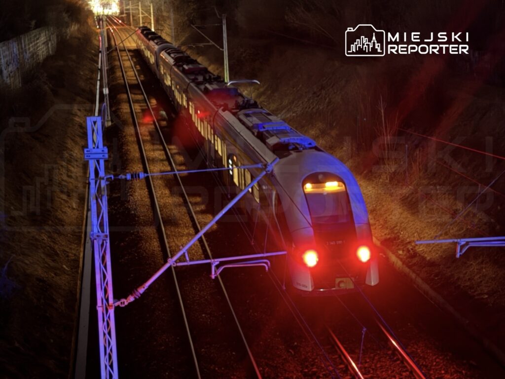 Pociąg osobowy z oświetleniem wjeżdża na stację kolejową w nocy, otoczony torami i ciemnym krajobrazem.