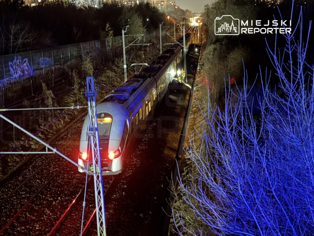 Pociąg osobowy zatrzymany na torach w nocy, obok widoczni pracownicy sprawdzający urządzenia, w tle miejskie oświetlenie.