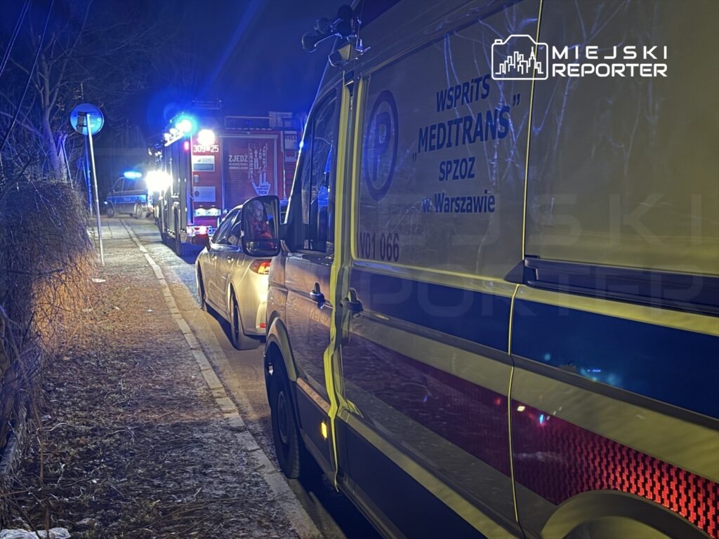 Karetka pogotowia i wóz strażacki zaparkowane na poboczu drogi, oświetlone niebieskimi i czerwonymi światłami.