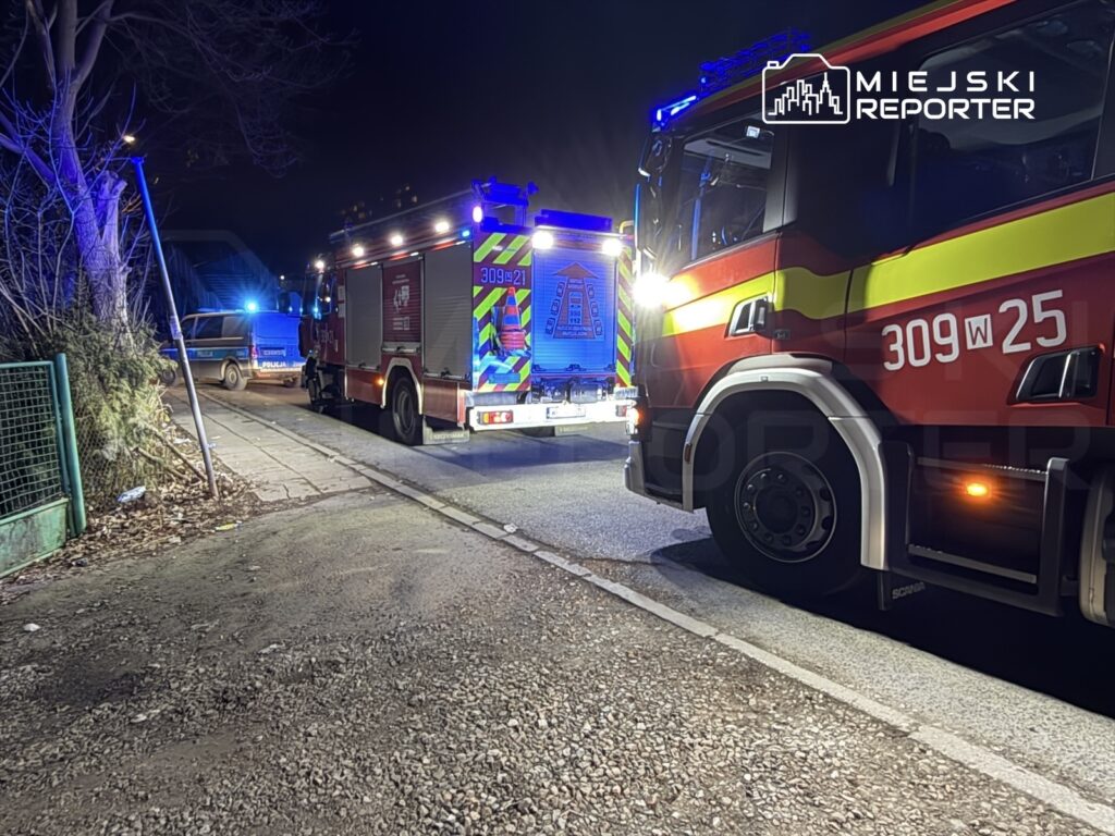 Czerwony wóz strażacki z włączonymi sygnałami świetlnymi stoi na ulicy obok policyjnego pojazdu, w nocy.