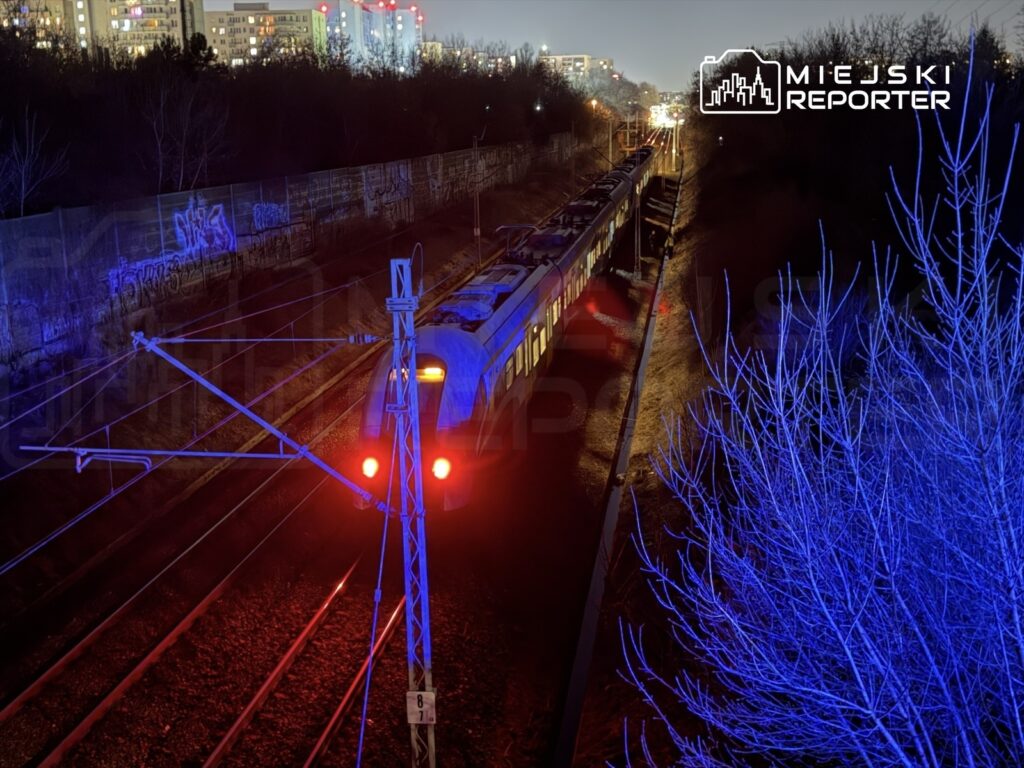 Pociąg osobowy zatrzymany na torach w nocy, oświetlony czerwonymi i niebieskimi światłami, w tle miejskie budynki.