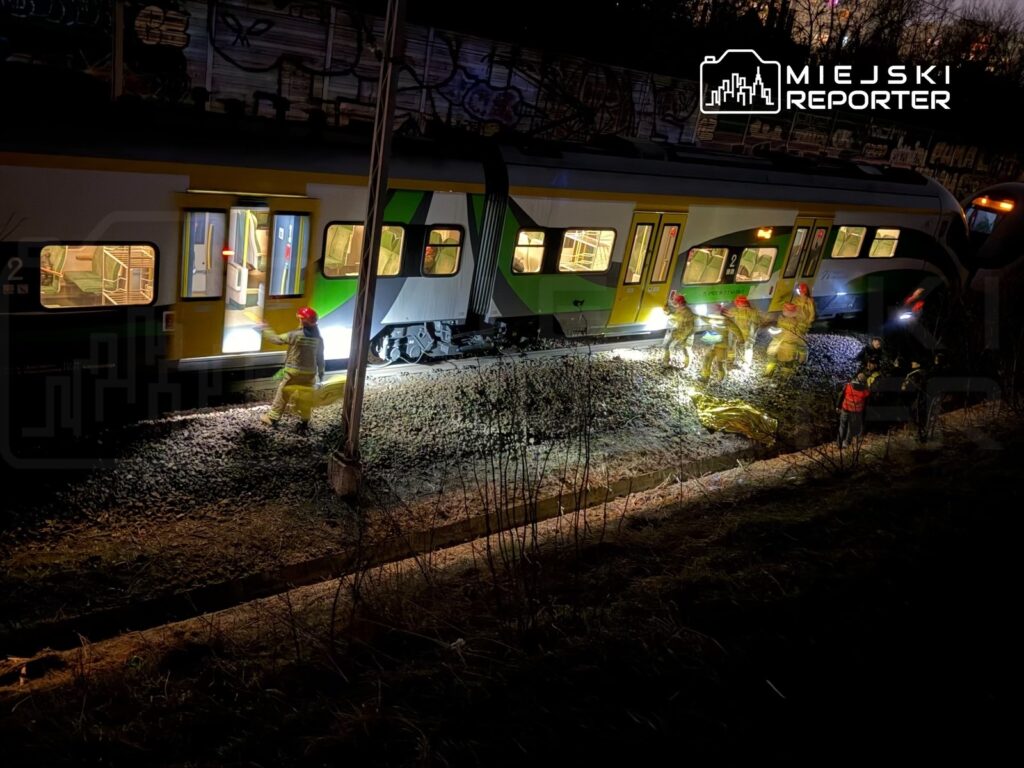 Grupa ratowników w hełmach prowadzi akcję ratunkową przy przewróconym pociągu w nocy, oświetlając teren latarkami.