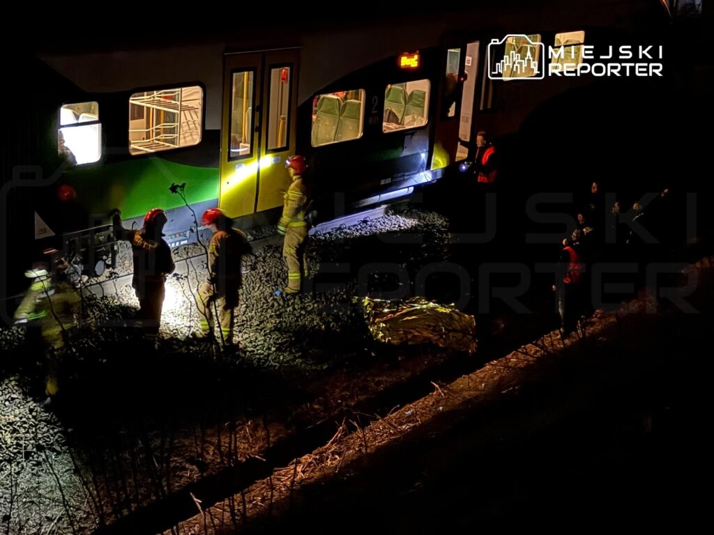 Grupa ratowników w hełmach pracuje przy torach kolejowych, obok stojącego pociągu, w nocy.