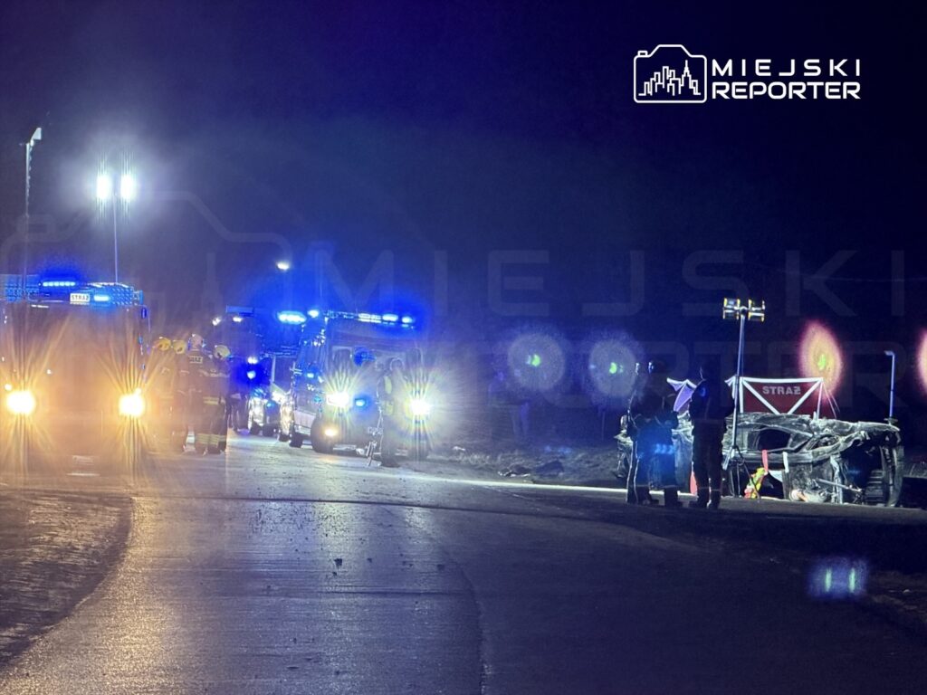 Grupa strażaków pracuje przy wypadku drogowym na oświetlonej drodze, obok przewróconego pojazdu.
