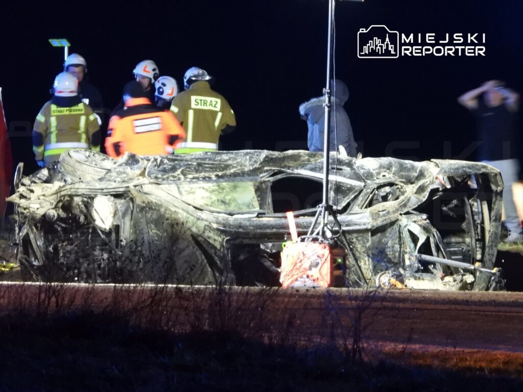 Zniszczony samochód leży na poboczu drogi, obok grupy ratowników w odzieży ochronnej.