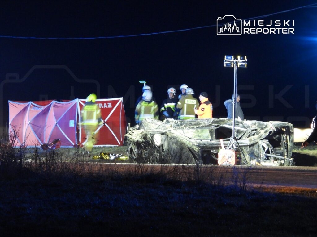 Ratownicy w mundurach przeszukują miejsce wypadku drogowego obok przewróconego samochodu w nocy.