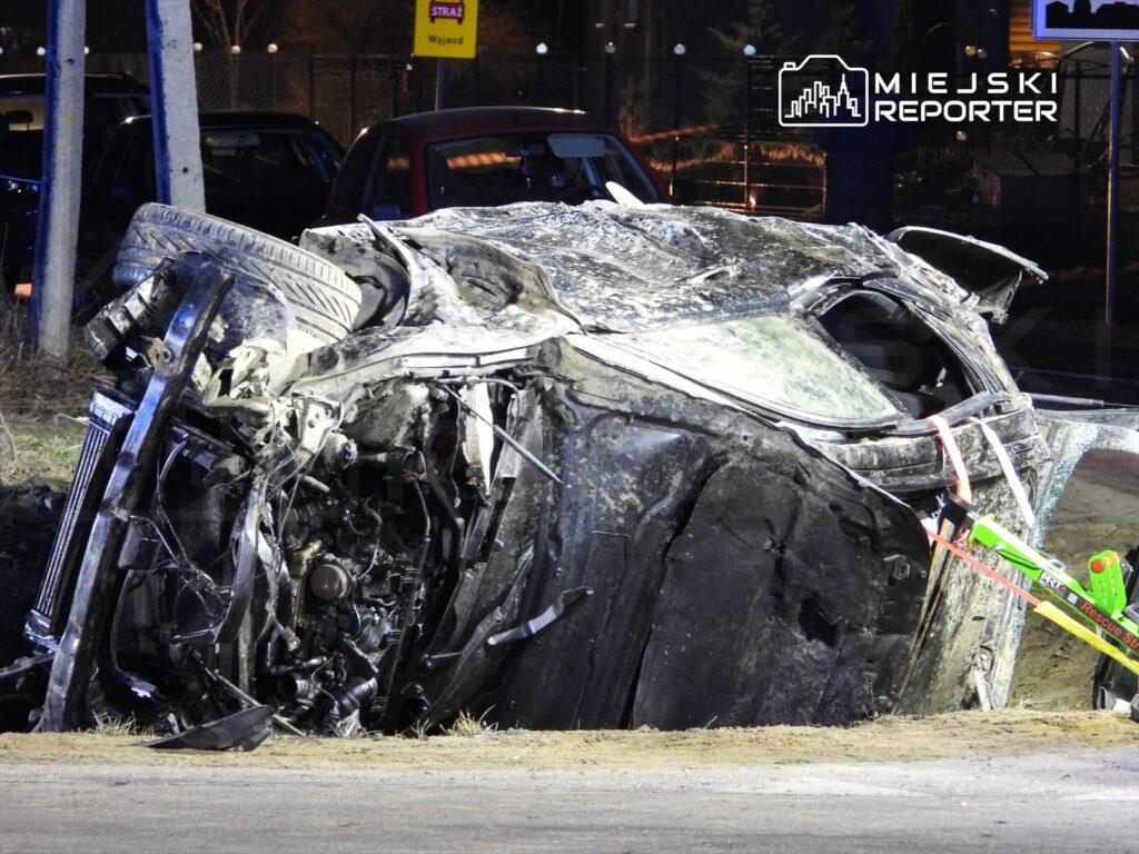 Wrak samochodu osobowego leży na dachu przy drodze, obok widać oznaczenie "STRAŻ" oraz inne pojazdy.
