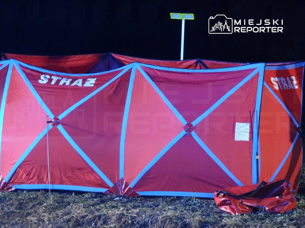 Czerwony namiot z napisem "STRAŻ" rozstawiony na trawie w ciemnym otoczeniu, oświetlony lampą.