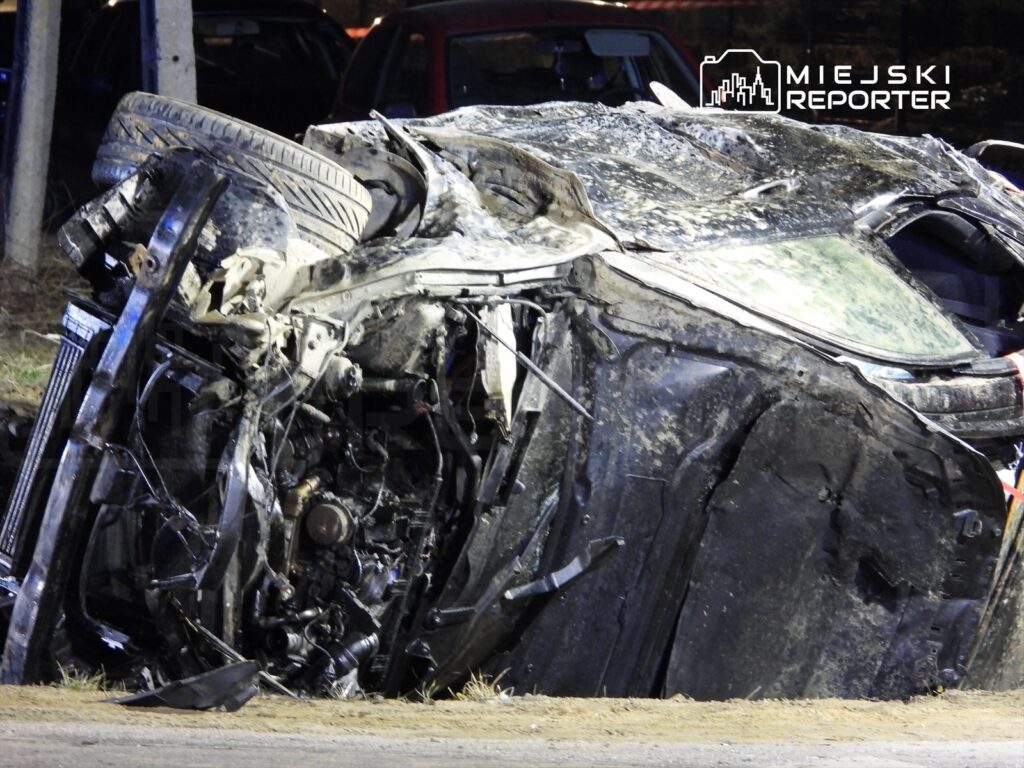 Zniszczony samochód leży na boku na poboczu drogi, z widocznymi uszkodzeniami nadwozia i silnika.