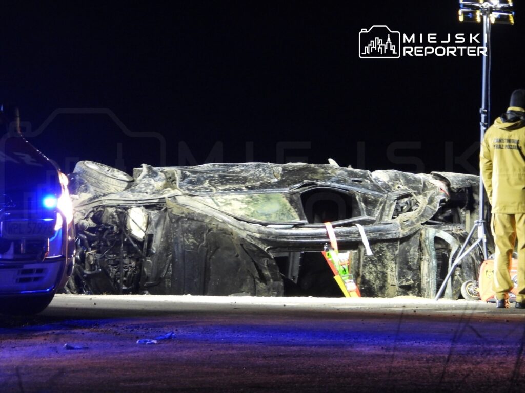 Przewrócony samochód osobowy leży na drodze, otoczony przez policyjny radiowóz z włączonymi światłami.