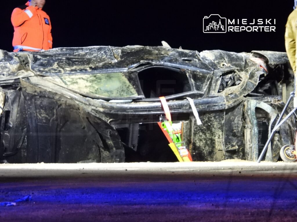 Mężczyzna w pomarańczowej kurtce rozmawia przez telefon obok przewróconego samochodu na drodze nocą.