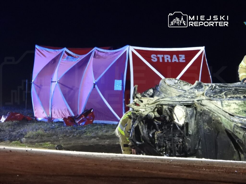 Strażacy pracują przy przewróconym pojeździe na drodze, obok rozstawiony namiot ratunkowy z napisem „STRAŻ”.