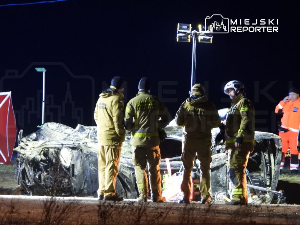 Grupa ratowników w specjalnych strojach omawia sytuację przy przewróconym samochodzie na drodze w nocy.