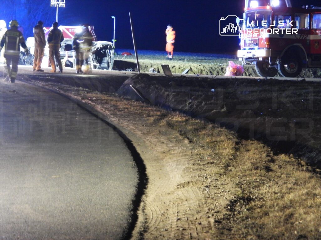 Grupa strażaków pracuje przy przewróconym pojeździe na drodze w nocy, w tle widać woz strażacki i oświetlenie.