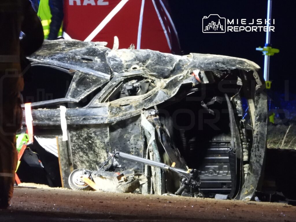 Wrak samochodu leży na boku na drodze, otoczony przez osoby w odblaskowych kamizelkach, w nocy.