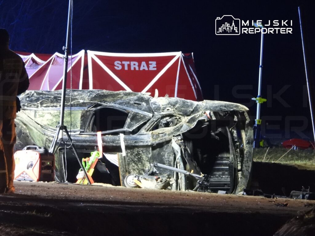 W nocy strażacy pracują przy przewróconym samochodzie na poboczu drogi, obok stoi namiot ratunkowy.