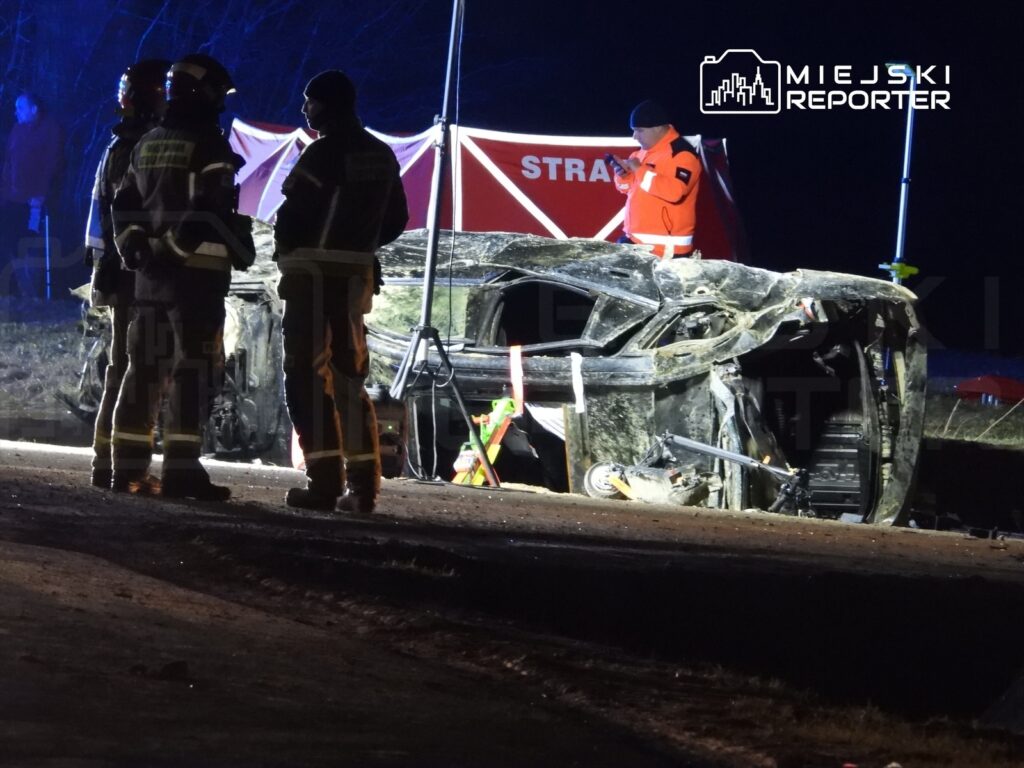 Grupa strażaków w mundurach omawia sytuację przy przewróconym samochodzie na drodze, w tle widoczna lampka ostrzegawcza.