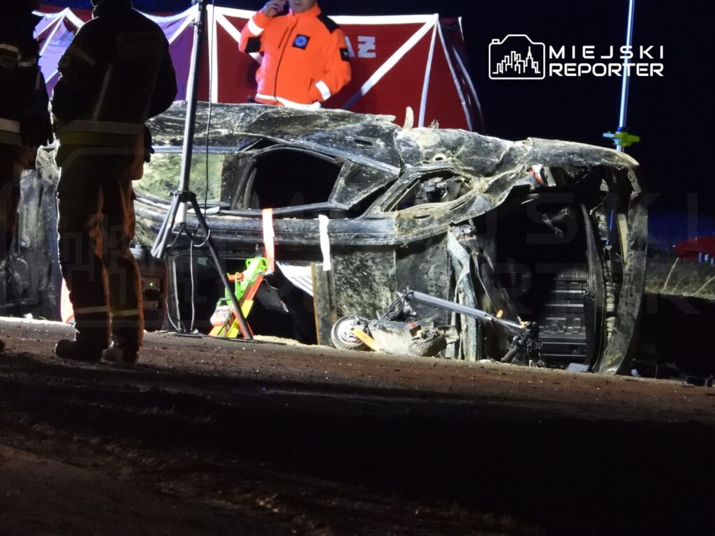 Zniszczony samochód leży na boku na drodze, a obok pracują strażacy w mundurach.