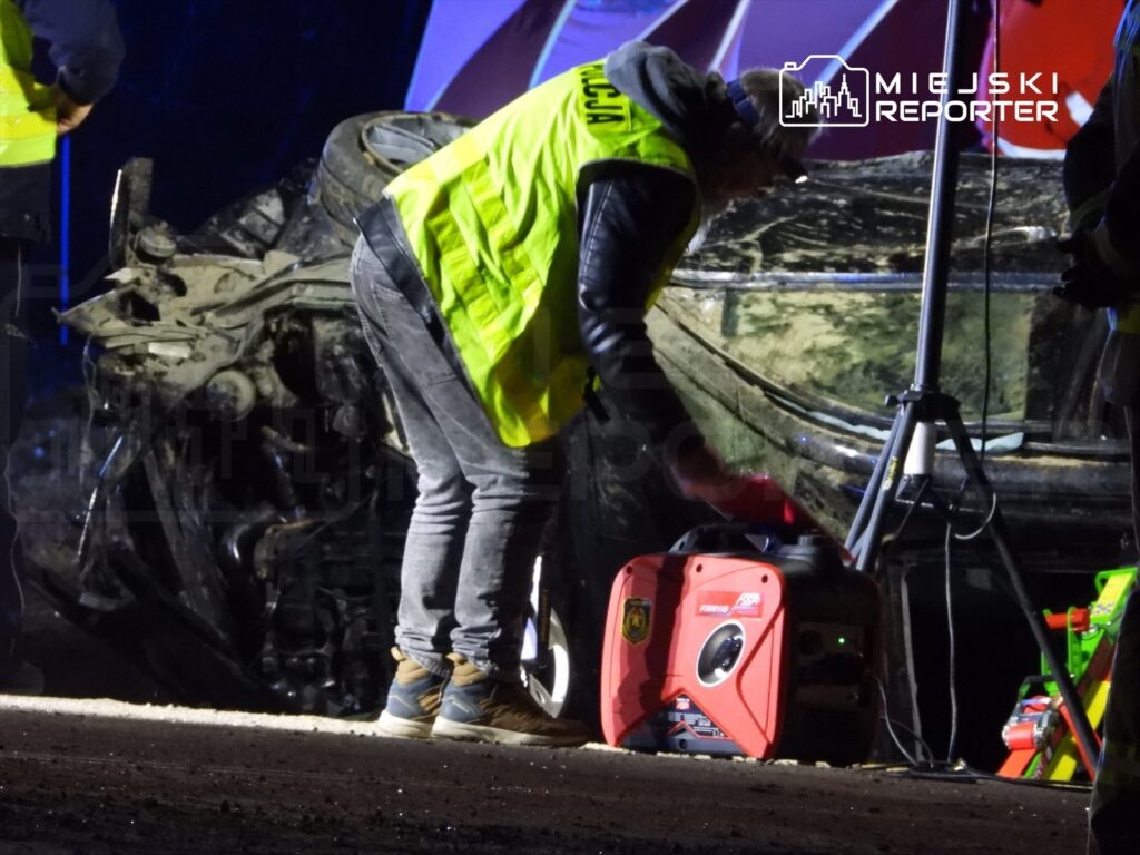 Grupa ratowników w odblaskowych kamizelkach pracuje przy przewróconym samochodzie, przygotowując sprzęt ratunkowy.