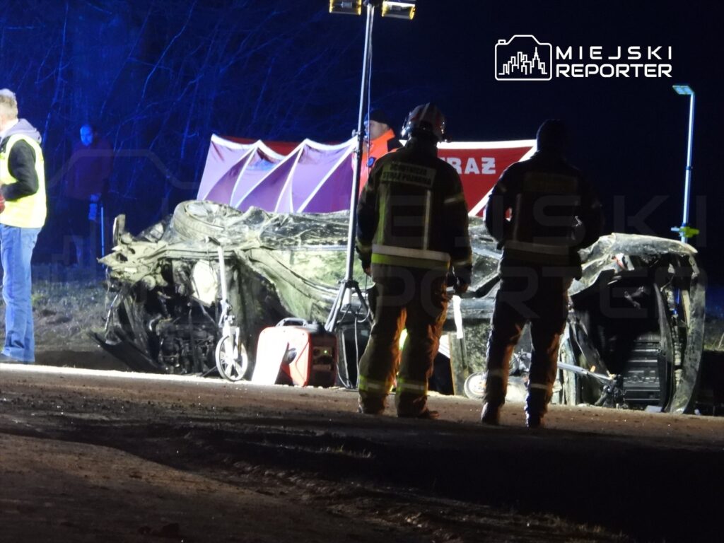 Na poboczu drogi leży zniszczony samochód, a obok niego stoją strażacy w mundurach, pracując przy akcji ratunkowej.